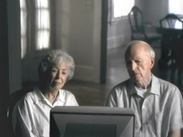 An elderly couple, a woman with white hair and a man with a receding hairline, sit side-by-side, looking intently at a computer monitor in a dimly lit room.