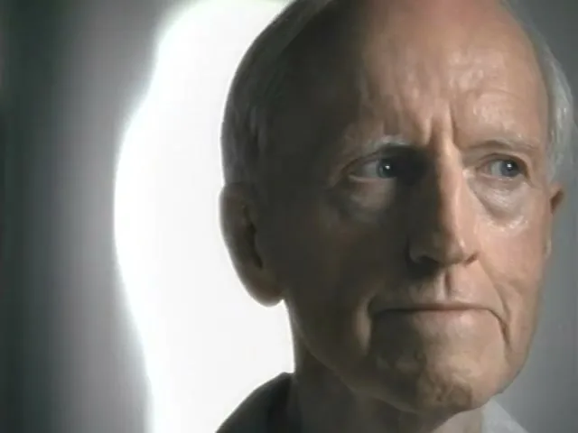 An elderly man with white hair and a light-colored shirt looks off-camera in a softly lit interior setting.