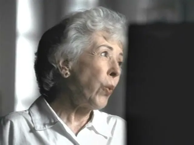 An elderly woman with grey hair and a white shirt looks up and to the right, making a surprised expression, in an interior setting.