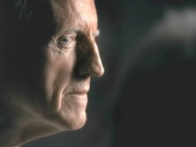 A man with a wrinkled face and blue eyes is shown in profile, looking off-screen with a neutral expression in a dimly lit interior.