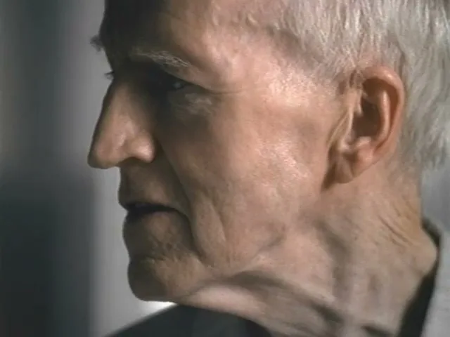 An elderly man with white hair and a wrinkled face looks off-screen, his mouth slightly open, in a medium closeup shot.