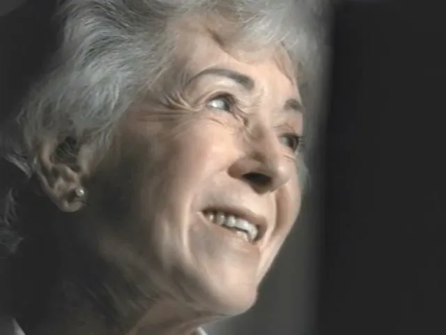 An elderly woman with white hair and a pearl earring smiles and looks up, in an interior setting.