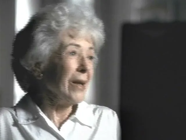 An elderly woman with white hair and a white shirt looks up and to the right, speaking in an interior setting.