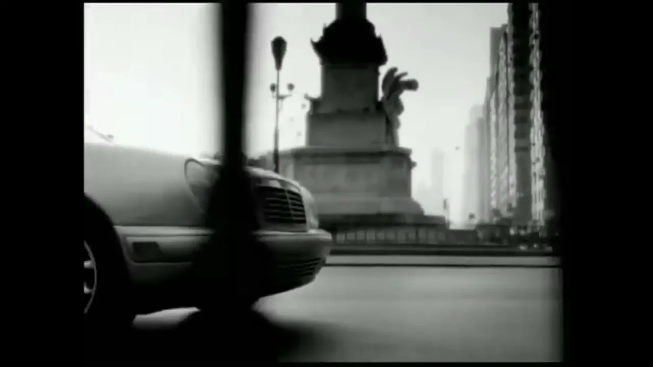 A Mercedes-Benz car drives on a city street, with a monument and buildings in the background, in black and white.