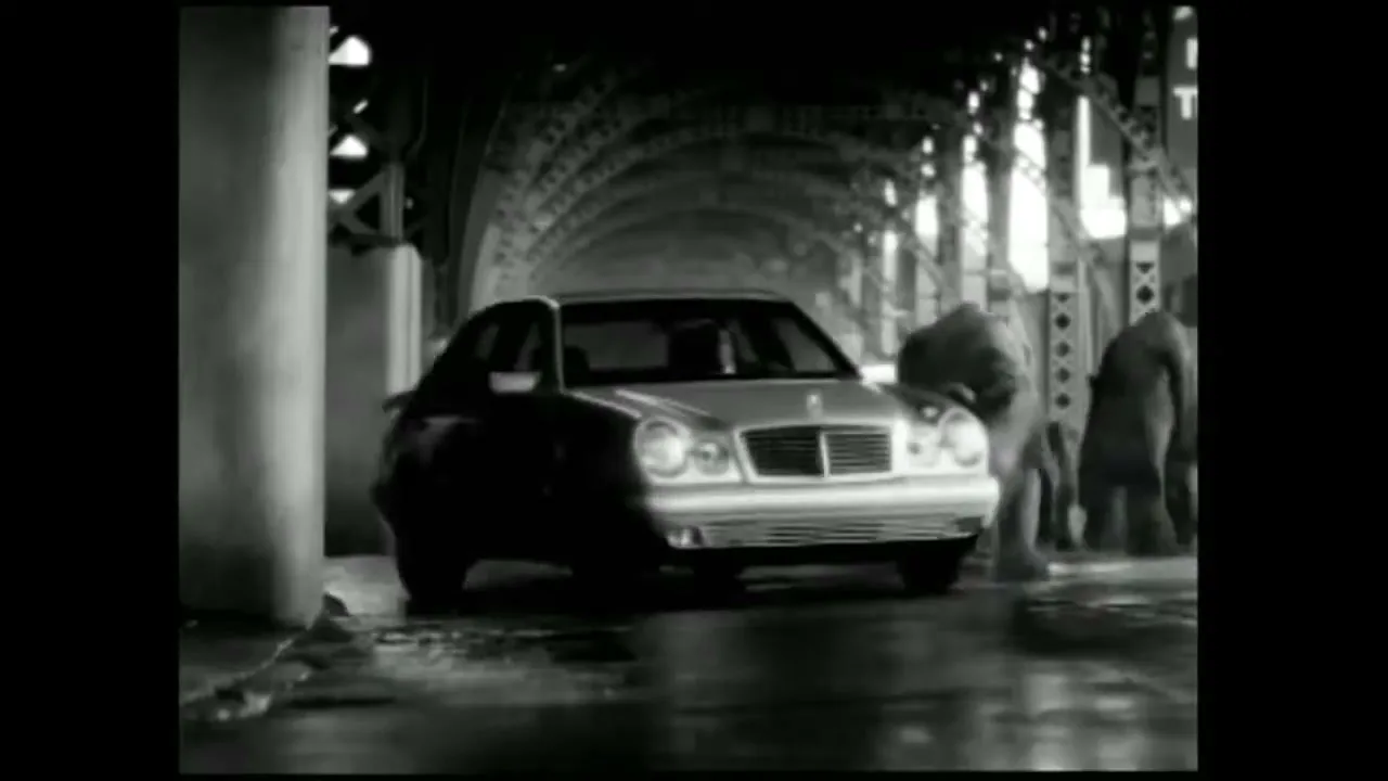 A silver Mercedes-Benz car drives through a dark, wet tunnel as rhinos walk past it in black and white.