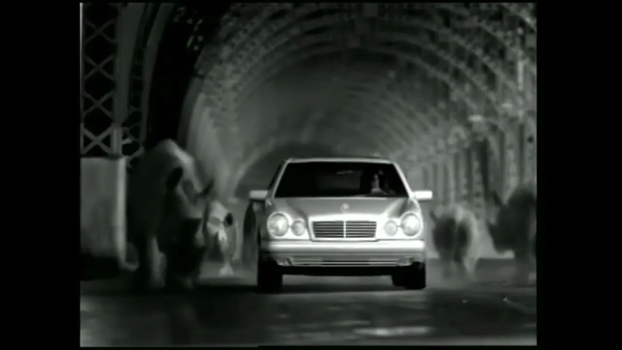 A silver Mercedes E-Class drives through a dark tunnel as a herd of rhinos stampedes around it, in black and white.