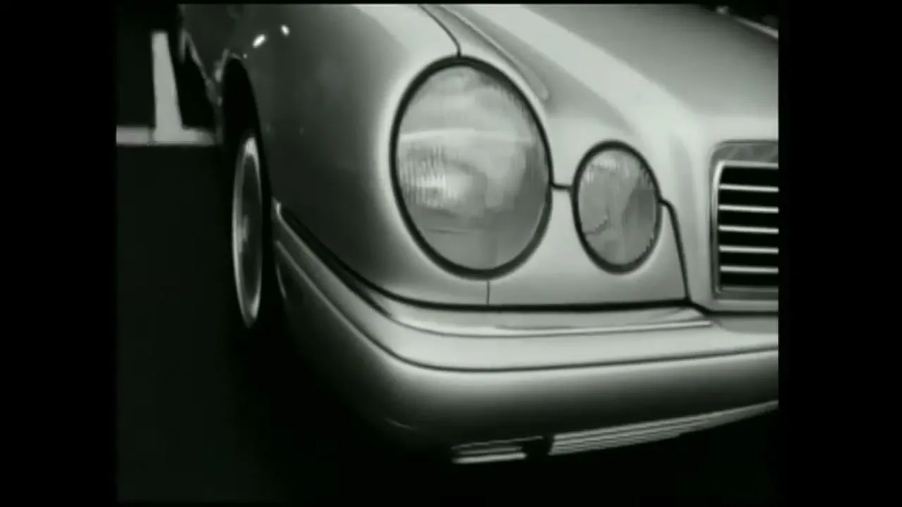 A close-up shot of a silver Mercedes-Benz car's front right headlight and grille, with the car moving forward on a dark surface.