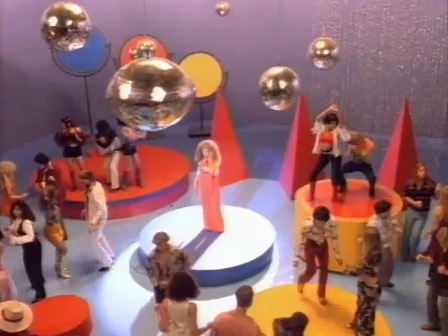 A female singer in a long orange dress stands on a circular stage while a crowd of people in 70s attire dance around her under disco balls.