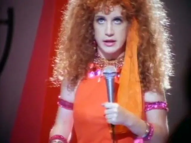 A woman with big curly red hair and an orange outfit sings into a microphone on a stage with red and white striped background.