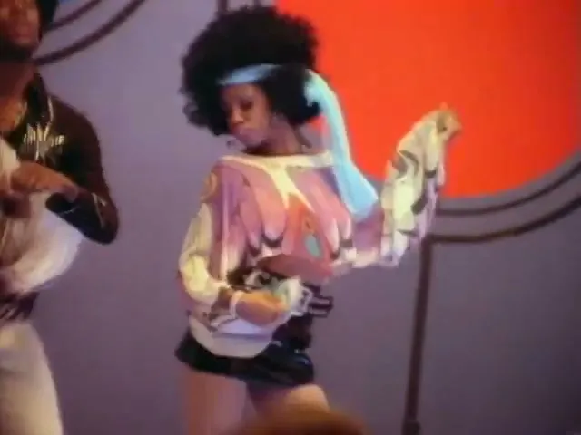 A man and a woman with afros and 70s attire dance on a stage with colorful circular backdrops.
