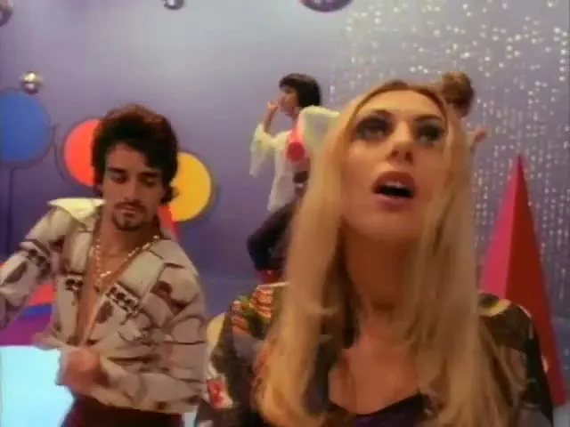 A blonde woman with heavy eye makeup and a patterned top looks up, then down, as she dances in a 70s-themed disco with other dancers.