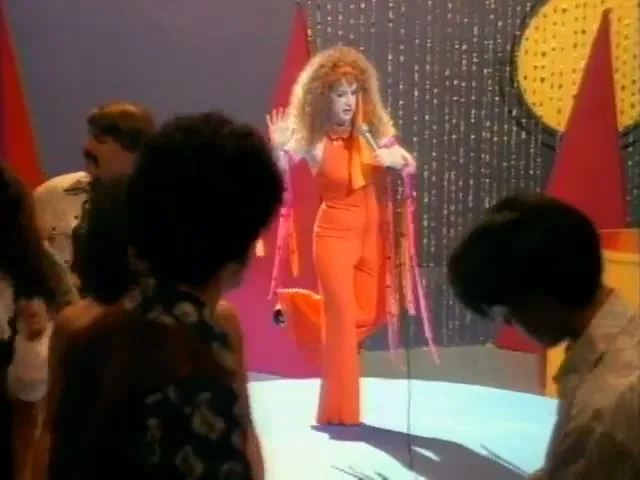 A woman with big curly hair in an orange jumpsuit sings into a microphone on a stage, surrounded by a small audience.