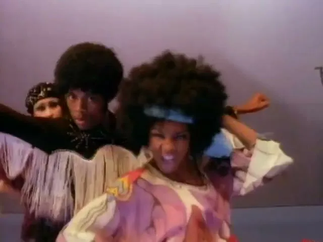 A group of three Black dancers with afros and 70s attire dance energetically in a studio void with a purple background.