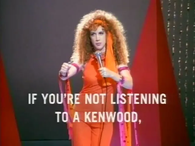 A woman with big curly red hair and an orange jumpsuit sings into a microphone on a stage with red and grey backdrops.