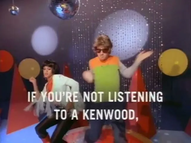 A man and a woman in 70s disco attire dance energetically in a brightly lit studio with disco balls and geometric shapes.