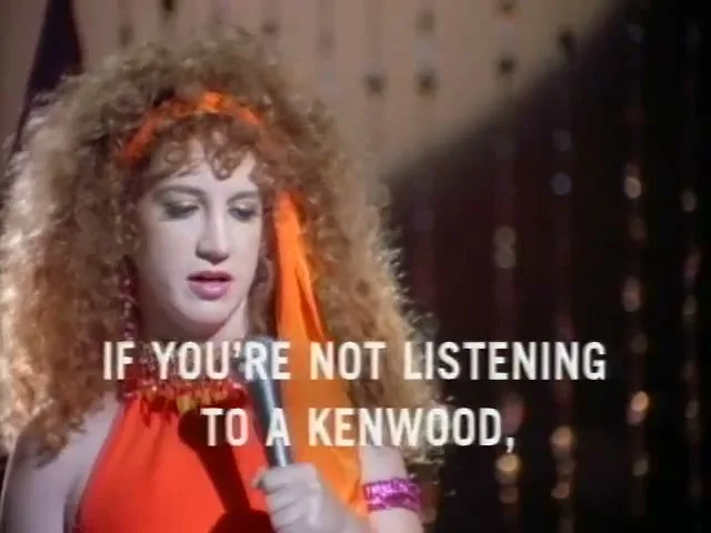A woman with big curly hair and an orange top sings into a microphone on a stage with text overlay.