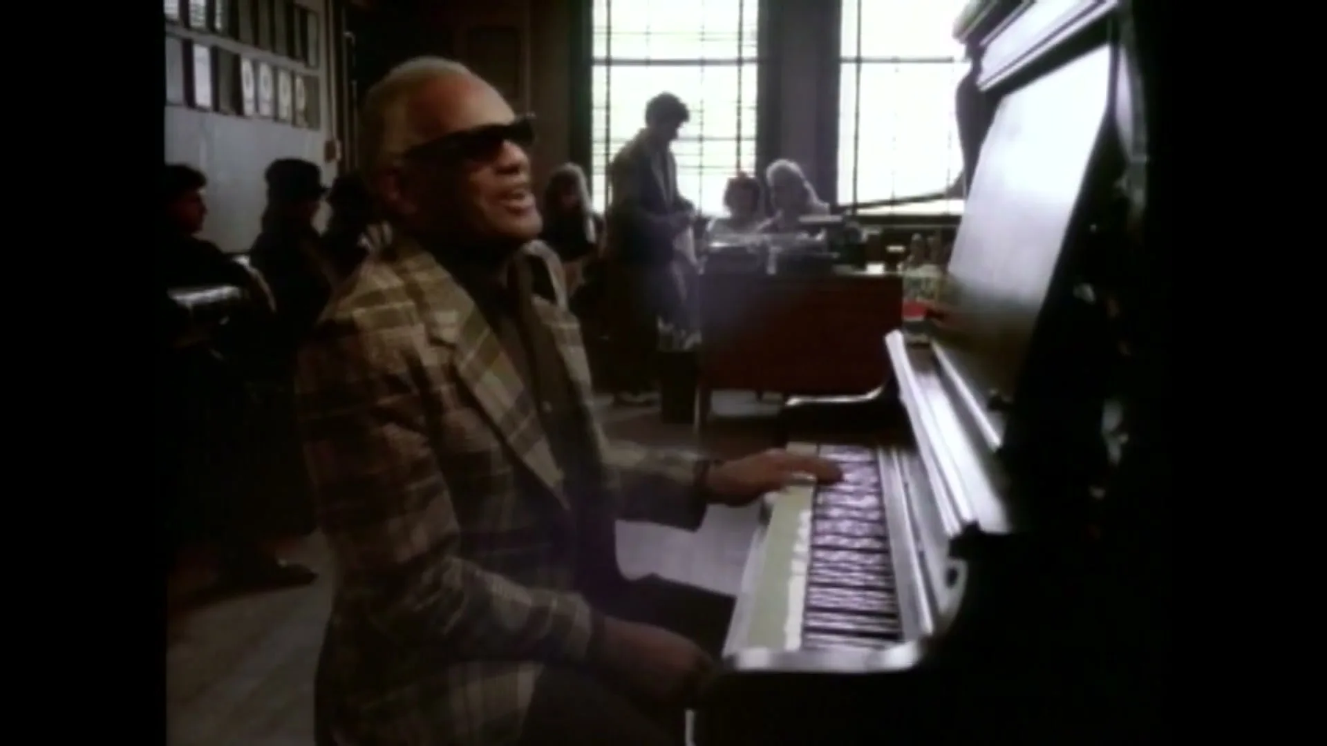 Ray Charles, a Black man in his 60s, wearing a plaid jacket and sunglasses, plays the piano and sings in a recording studio.