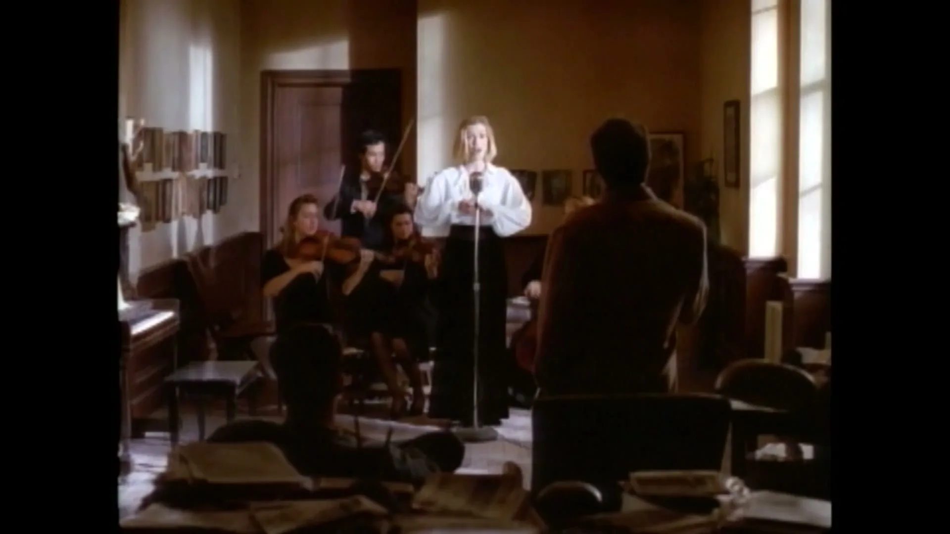 A female singer in a white blouse performs with a string quartet in a dimly lit room, while a man watches from the foreground.