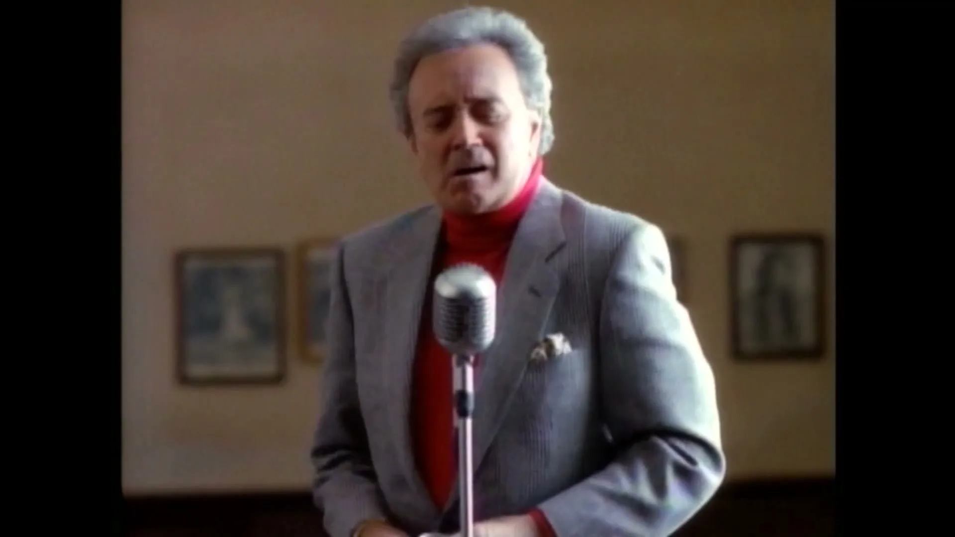 An older man with gray hair, wearing a gray suit and red turtleneck, sings into a vintage microphone in a room with framed pictures.