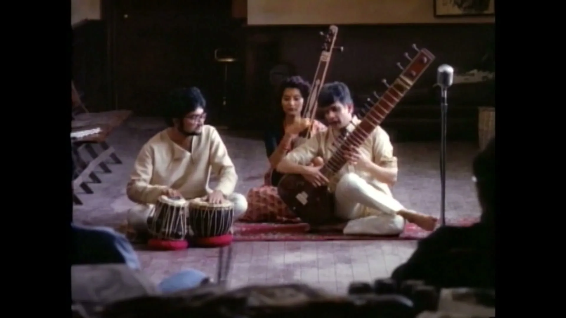 Three Indian musicians, two men and one woman, sit on a rug playing traditional instruments in a dimly lit room.