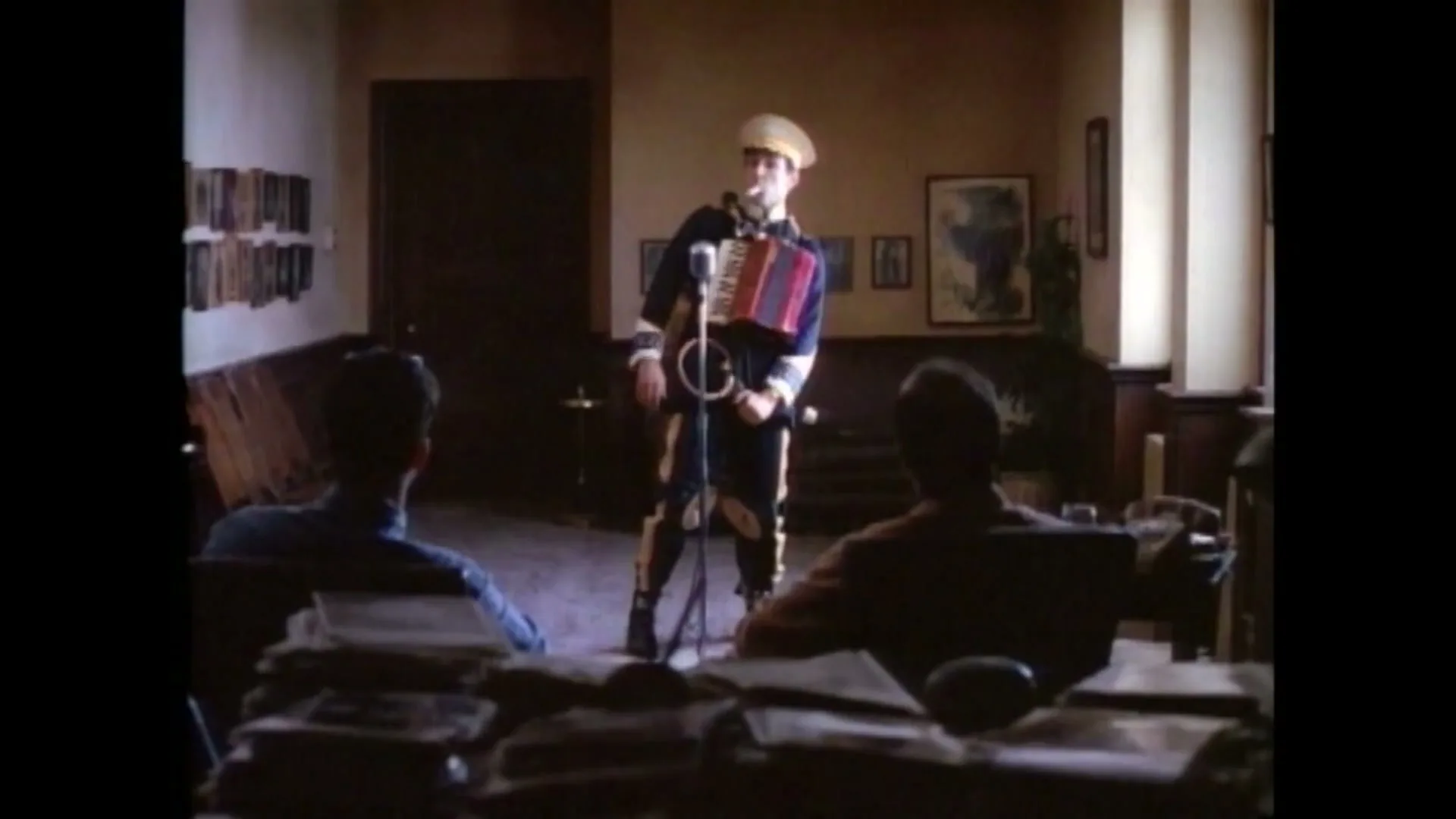 A man in a military-style uniform plays an accordion and sings into a microphone for two men sitting at a desk in a dimly lit office.