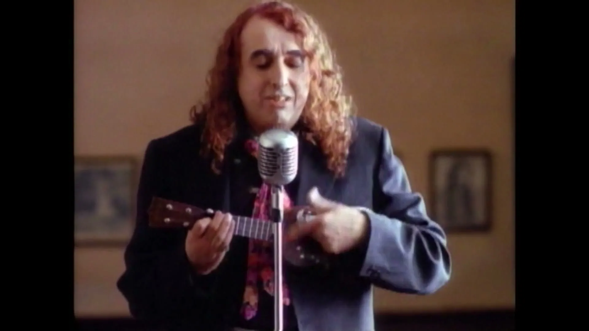 A man with long curly red hair and a black suit sings into a vintage microphone while playing a ukulele in a room with framed pictures.