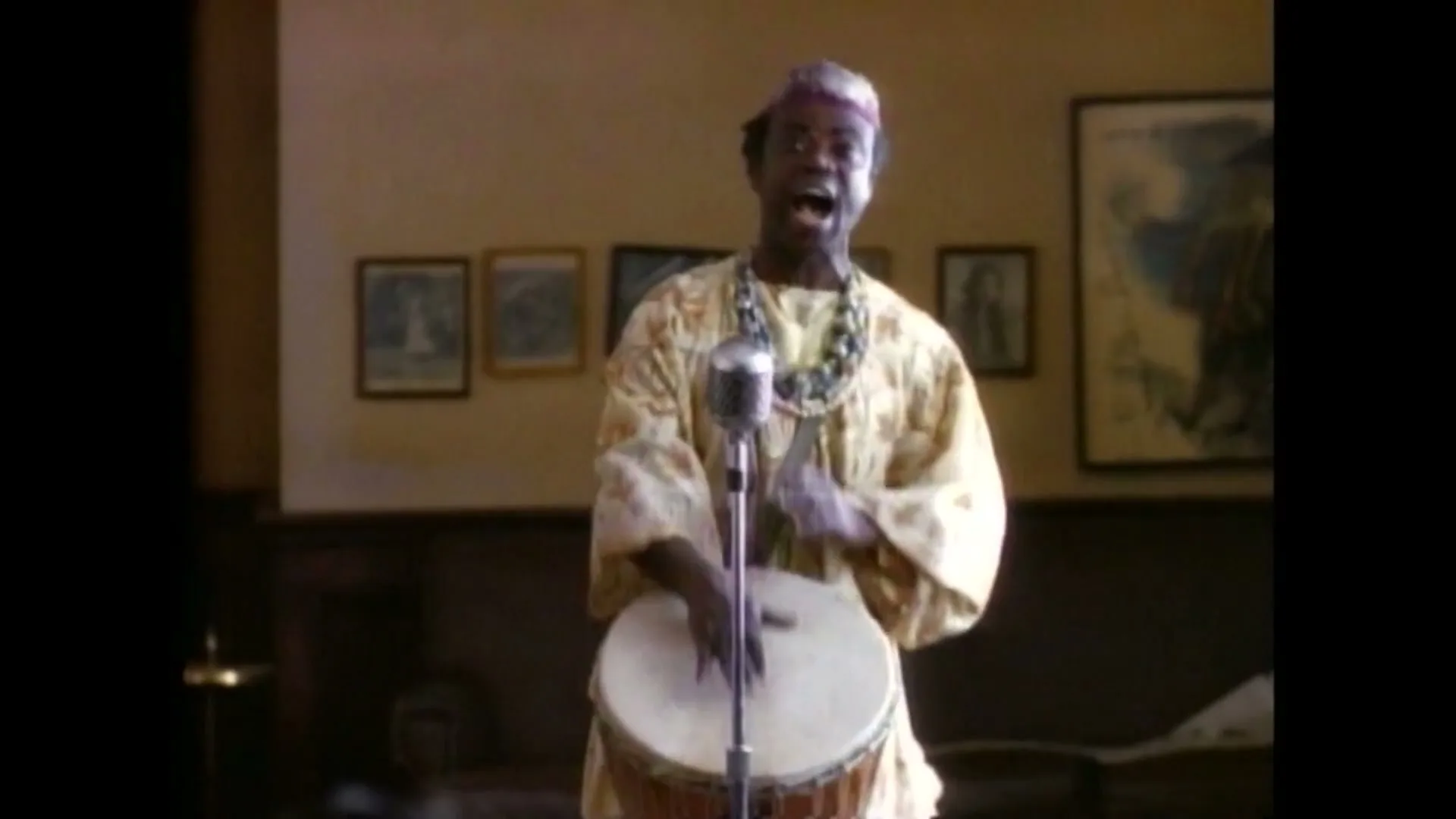 A man in traditional African attire plays a drum and sings into a vintage microphone during an audition in a room with framed pictures.