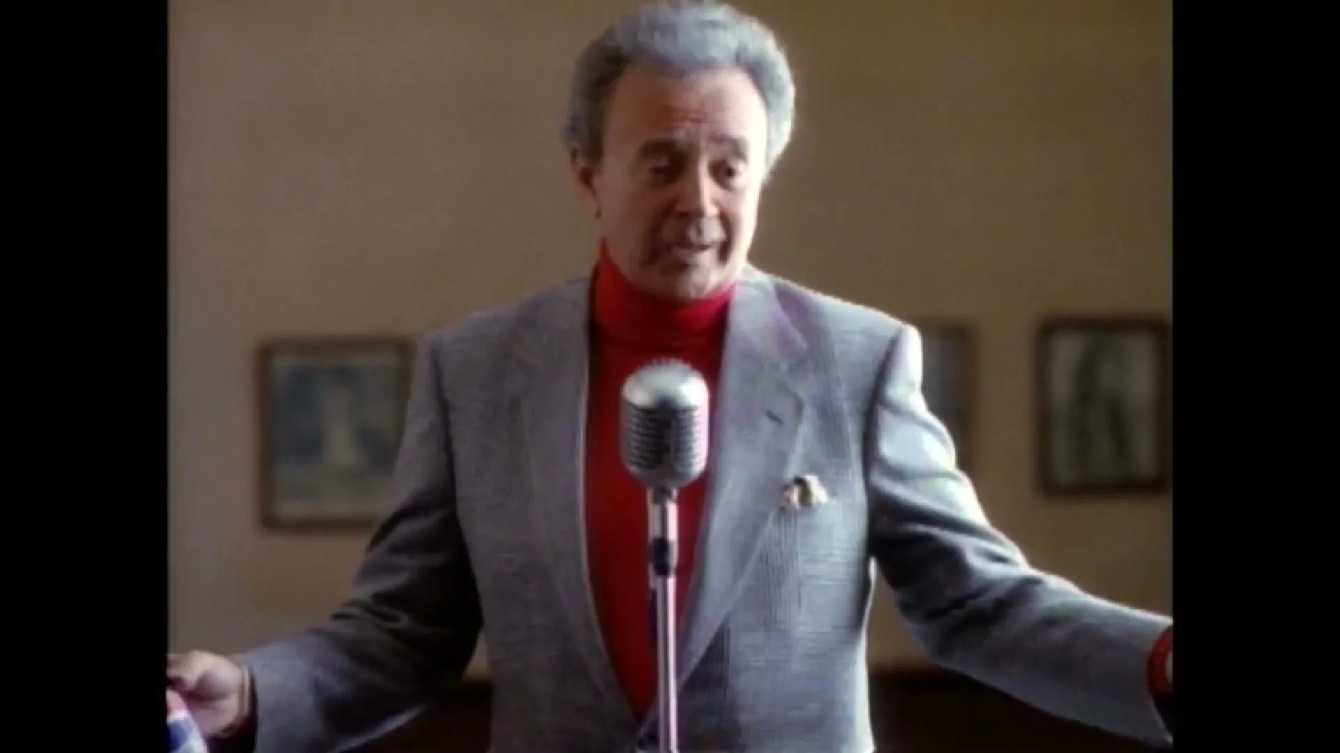 An older man with gray hair, wearing a gray suit and red turtleneck, stands in front of a microphone, singing in a room.