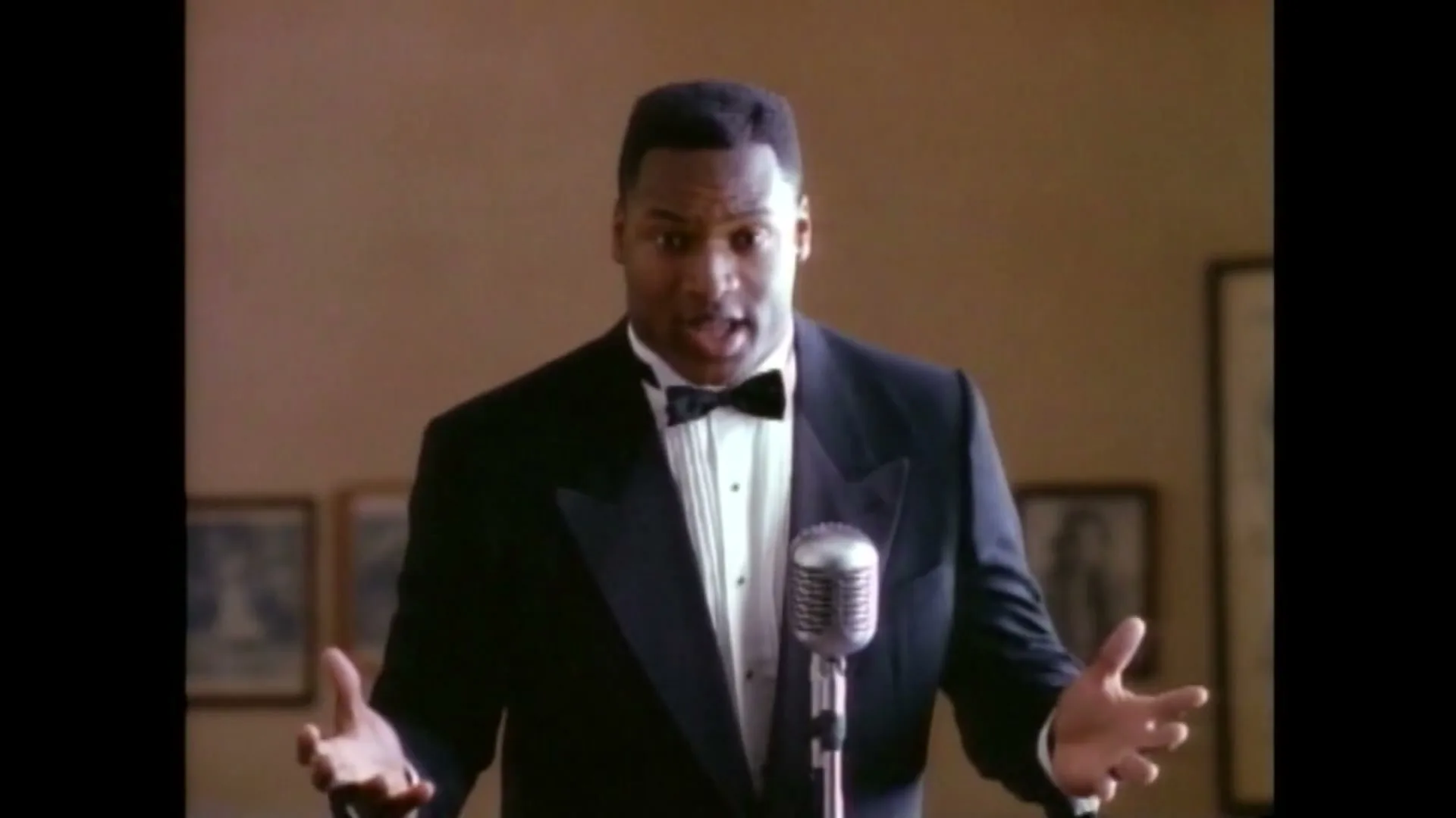 A Black man in his 30s, wearing a tuxedo, stands at a microphone with his arms outstretched, singing in an audition room.