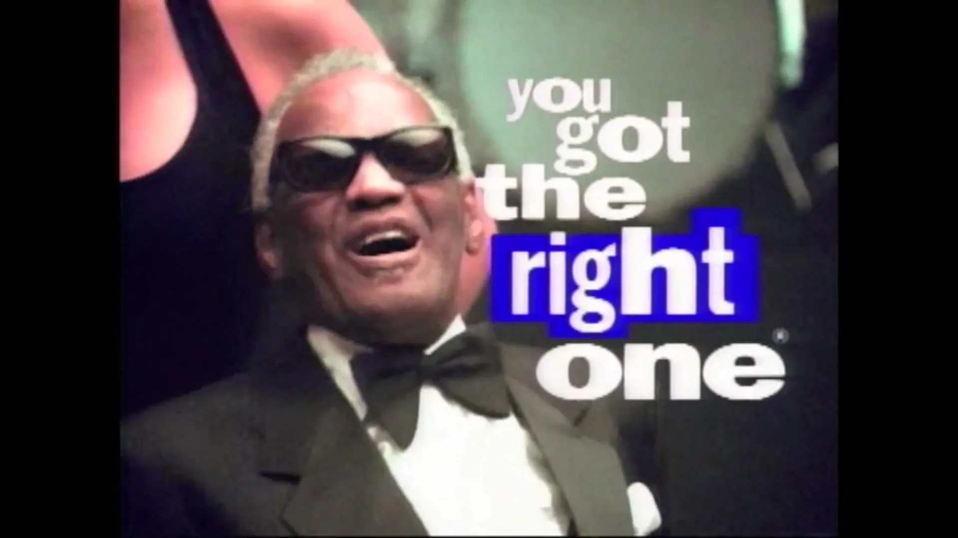 Ray Charles, a Black man in his 60s, wearing a tuxedo and sunglasses, sings with a joyful expression as a woman's arm is seen behind him, with text appearing on screen.