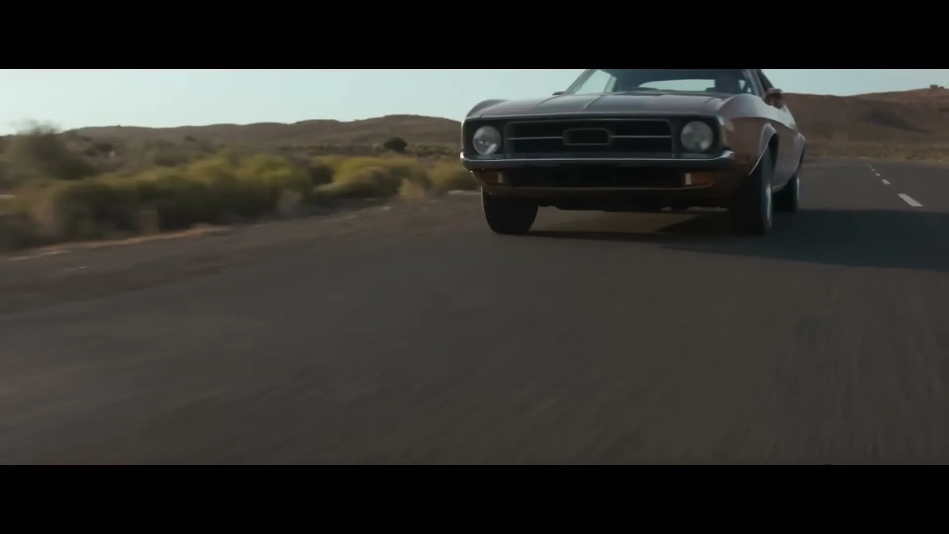 A vintage brown muscle car drives down a desert road, with a wide-angle shot capturing its movement and the arid landscape.