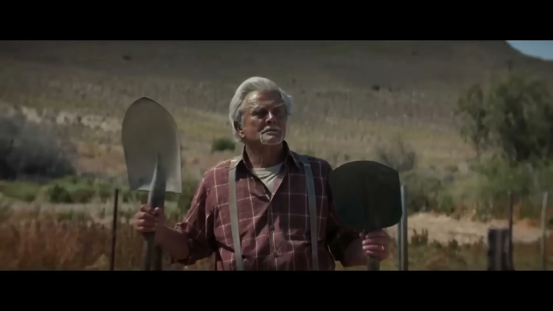 An older man with gray hair and a mustache stands in a dry, rural landscape, holding two shovels.