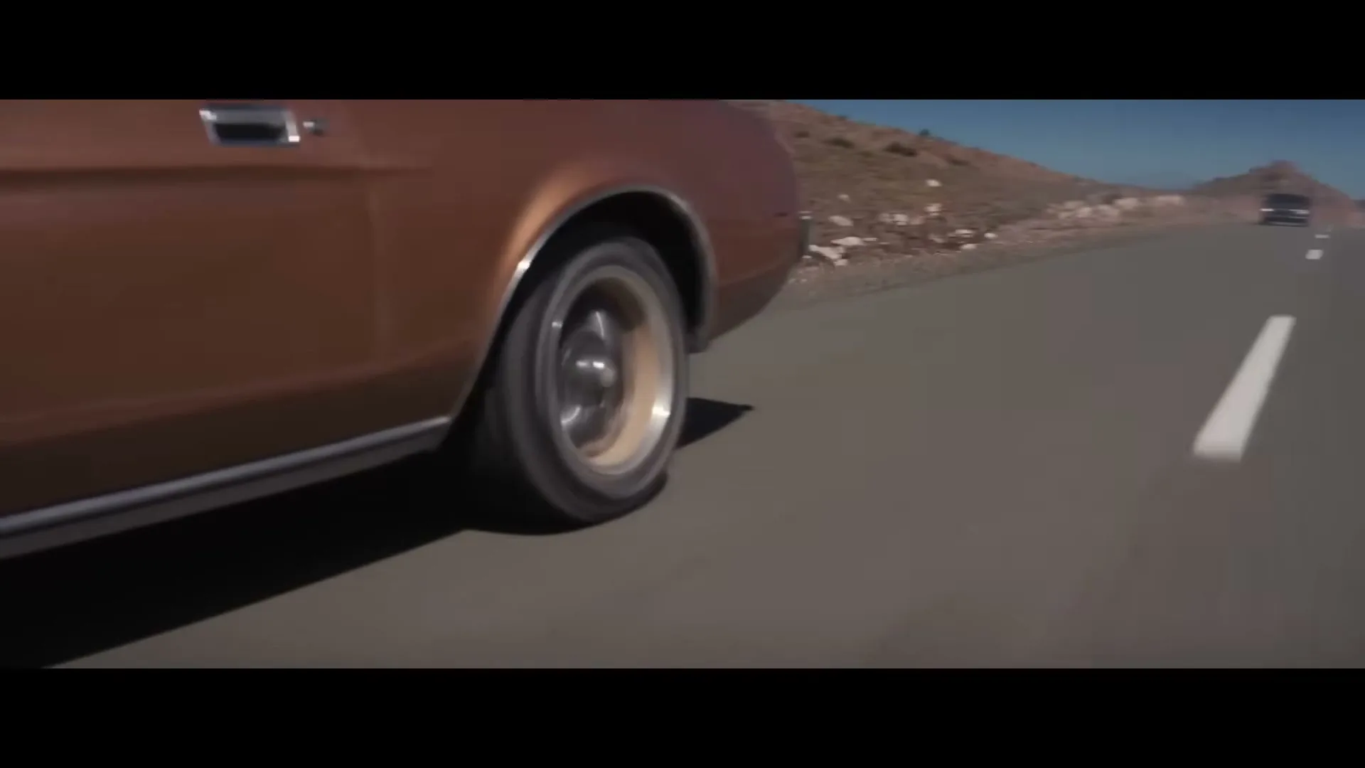 A brown vintage car drives on a desert road with mountains in the background under a clear blue sky.