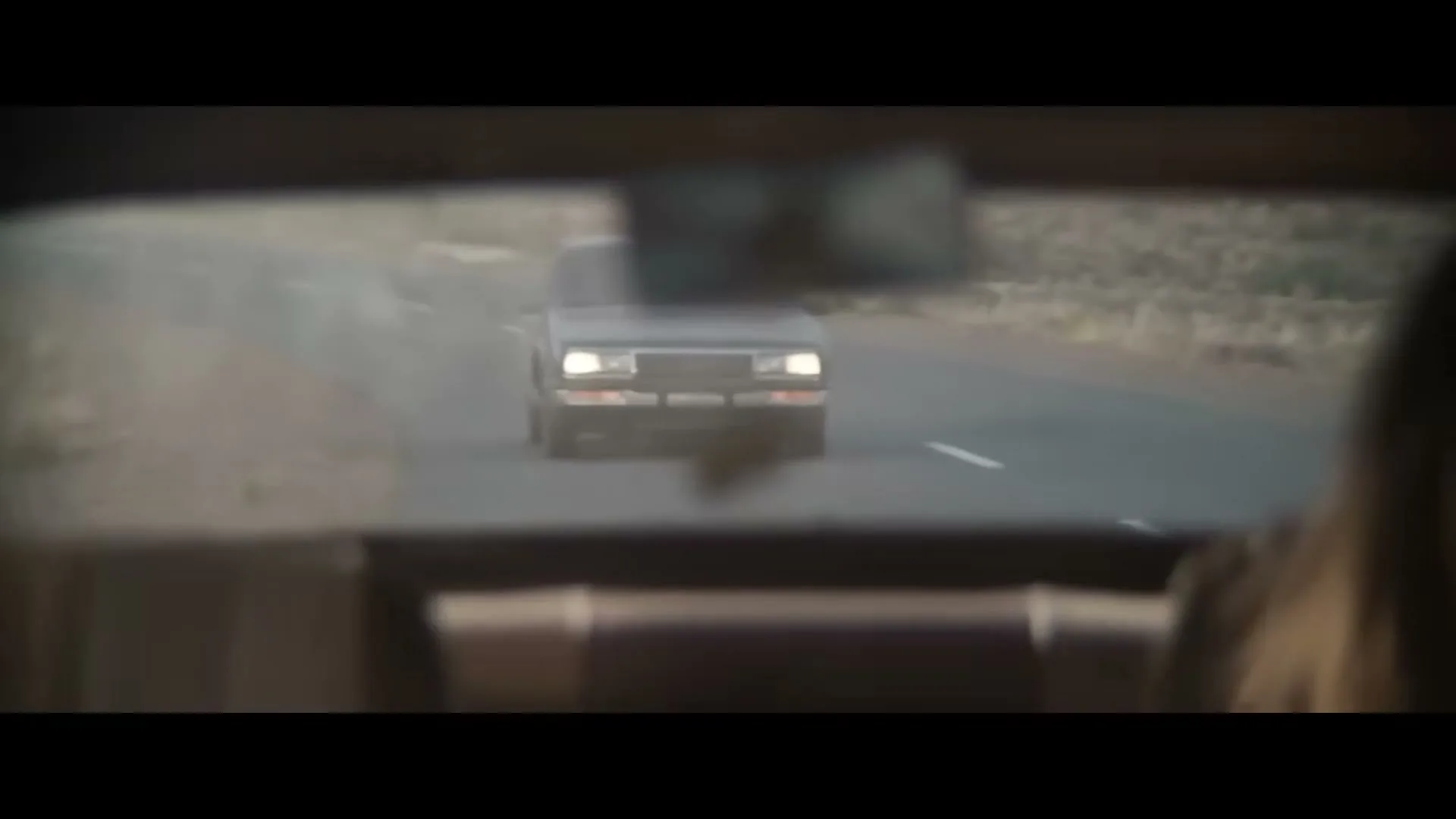 A man with long hair drives a car, looking in the rearview mirror at a truck approaching on a dusty road.