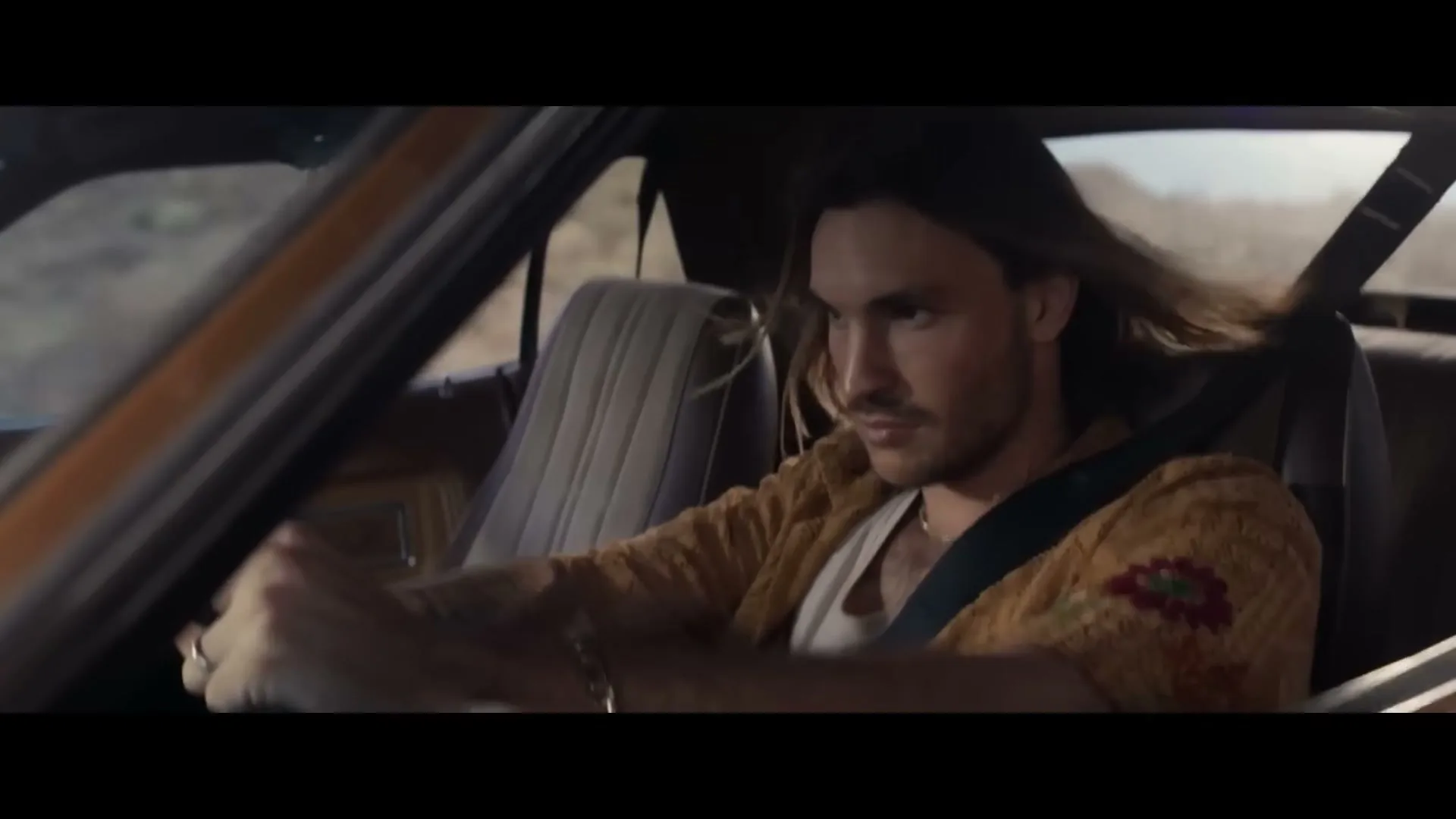 A man with long hair and a patterned cardigan drives a vintage car through a desert landscape, looking focused on the road.