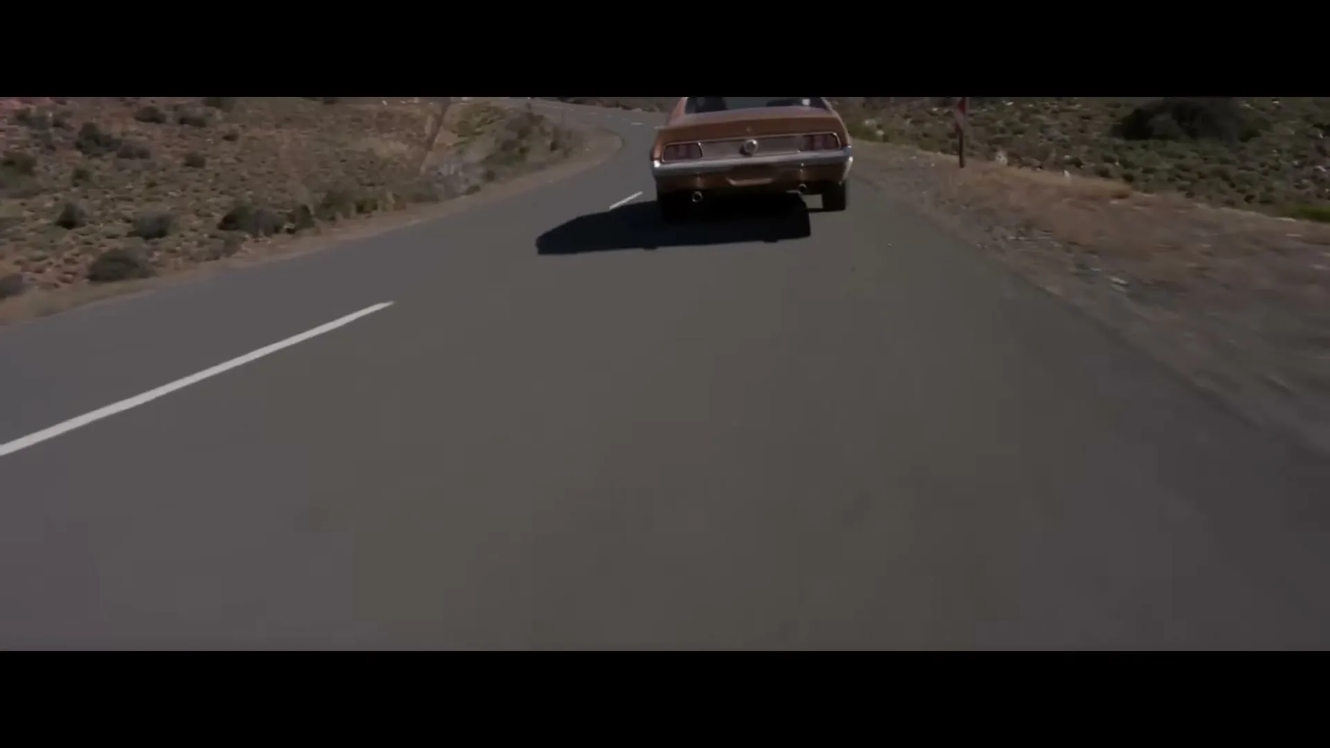A brown vintage car drives away from the camera on a winding desert road under a clear sky.
