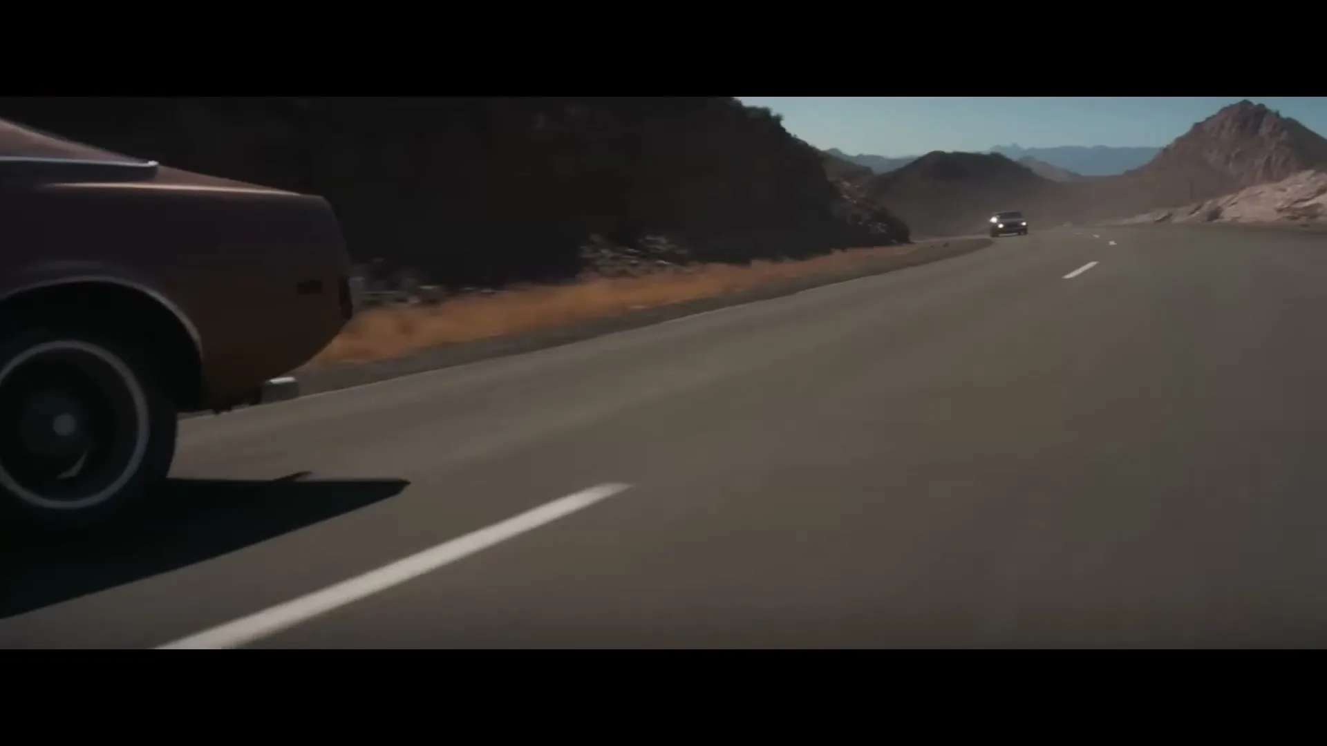 Two cars drive on a desert highway, one in the foreground and another in the distance, under a clear sky with mountains.