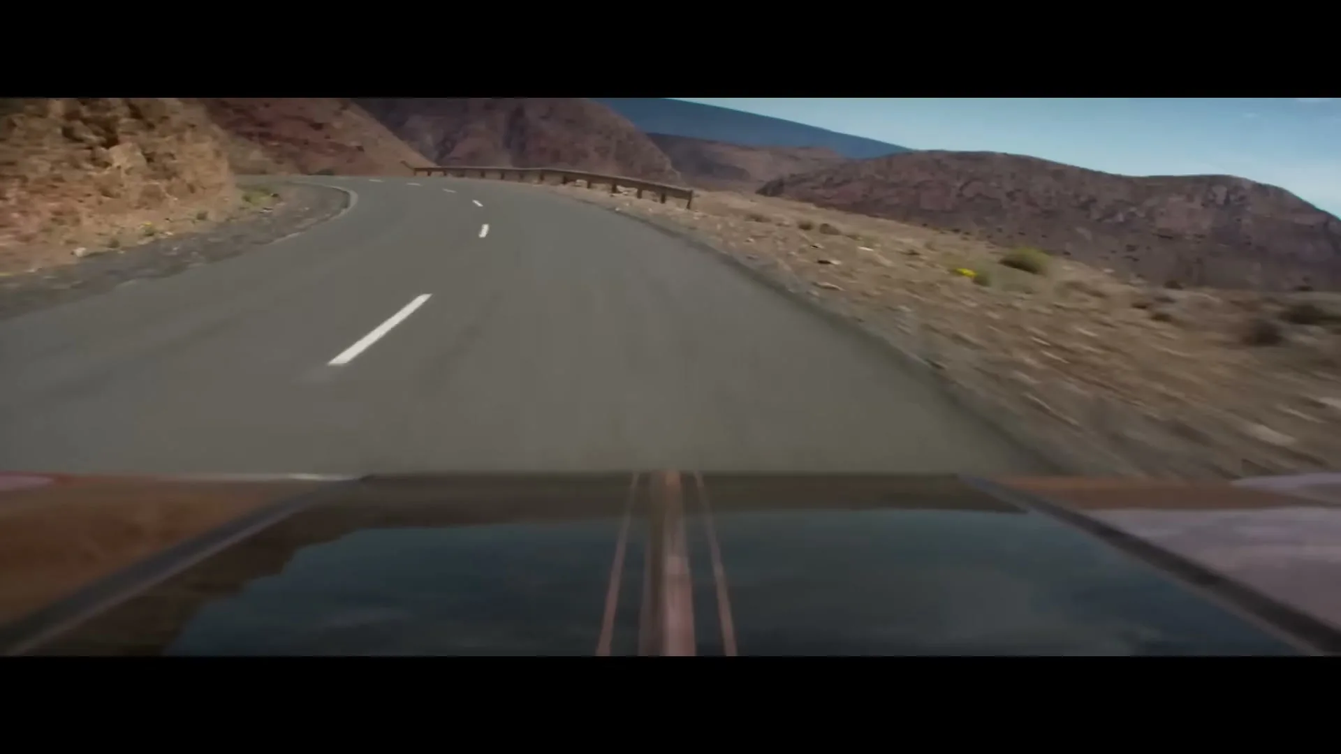 A car drives along a winding road through a mountainous desert landscape under a clear sky, with the car's hood visible in the foreground.