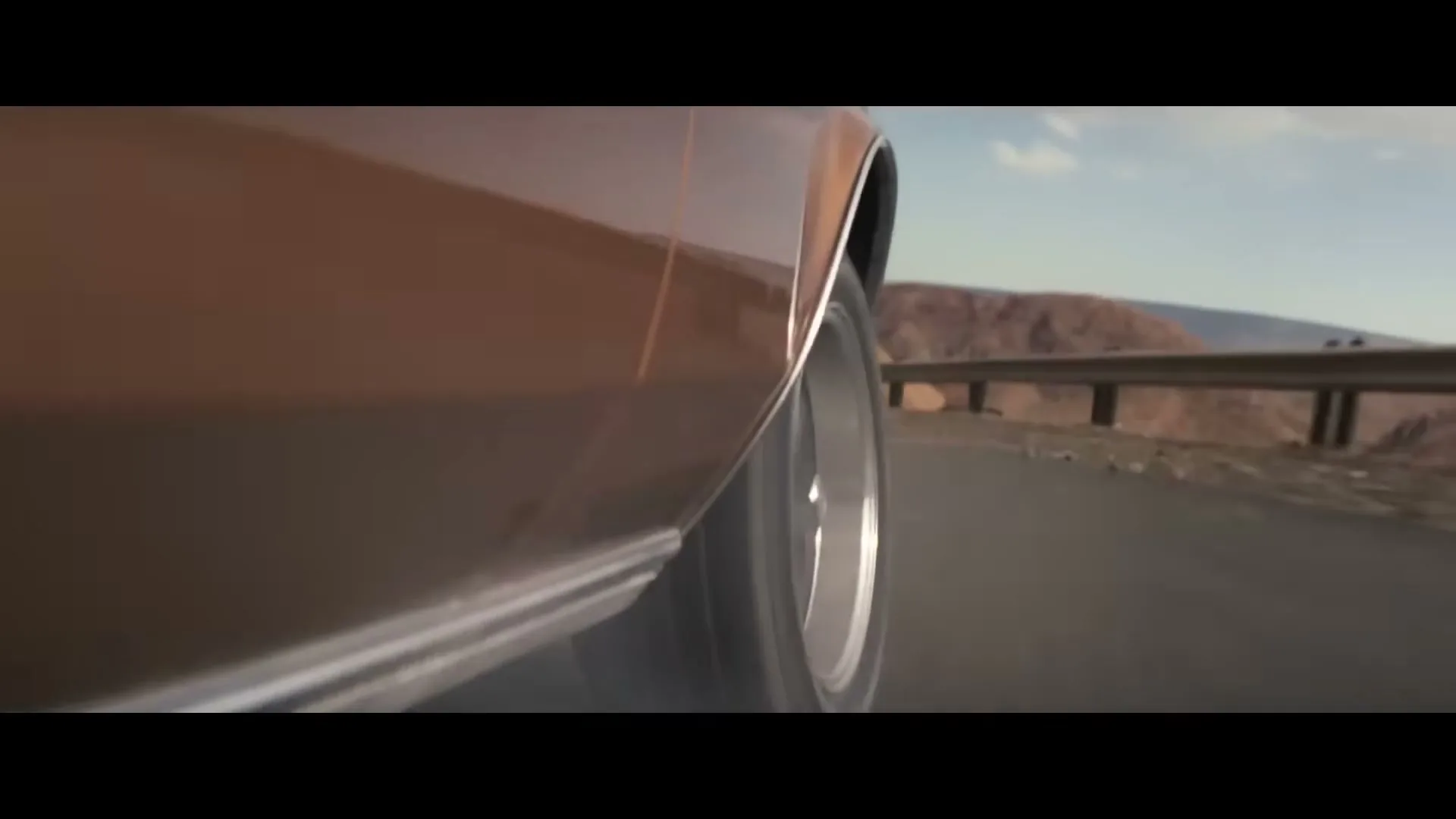 A close-up of a brown car's wheel and side as it drives along a winding road with mountains in the background.