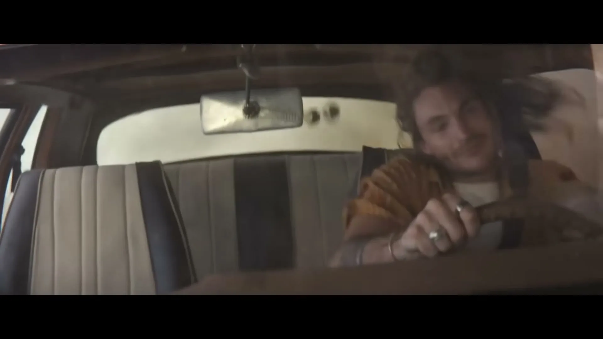 A young man with dreadlocks and a beard drives a car, looking happy and energetic as his hair flies around.
