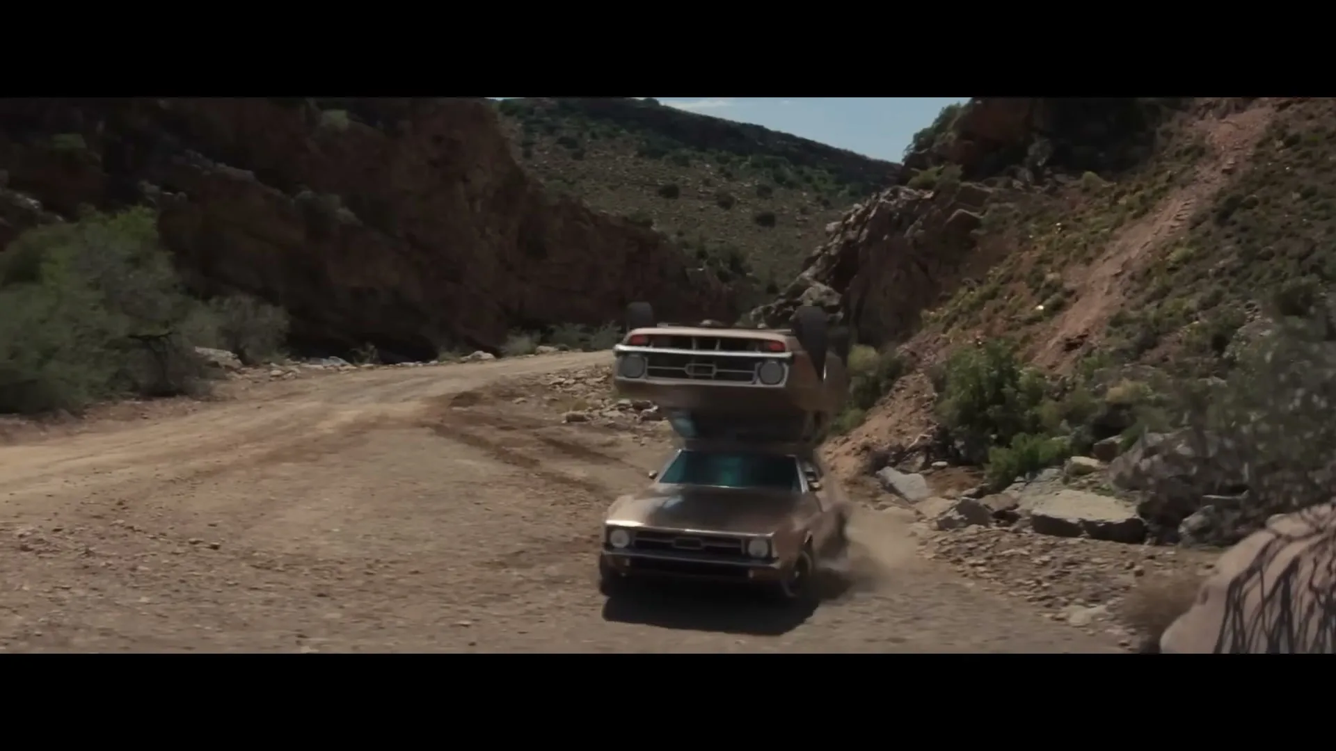 Two brown cars, one upside down on top of the other, speed down a dusty dirt road in a canyon on a sunny day.