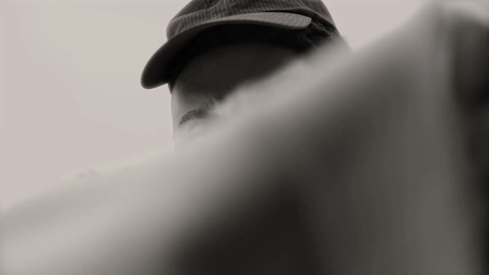 A black and white shot of a man with dreadlocks and a cap, looking directly at the camera, as a cloth is pulled away from his face.
