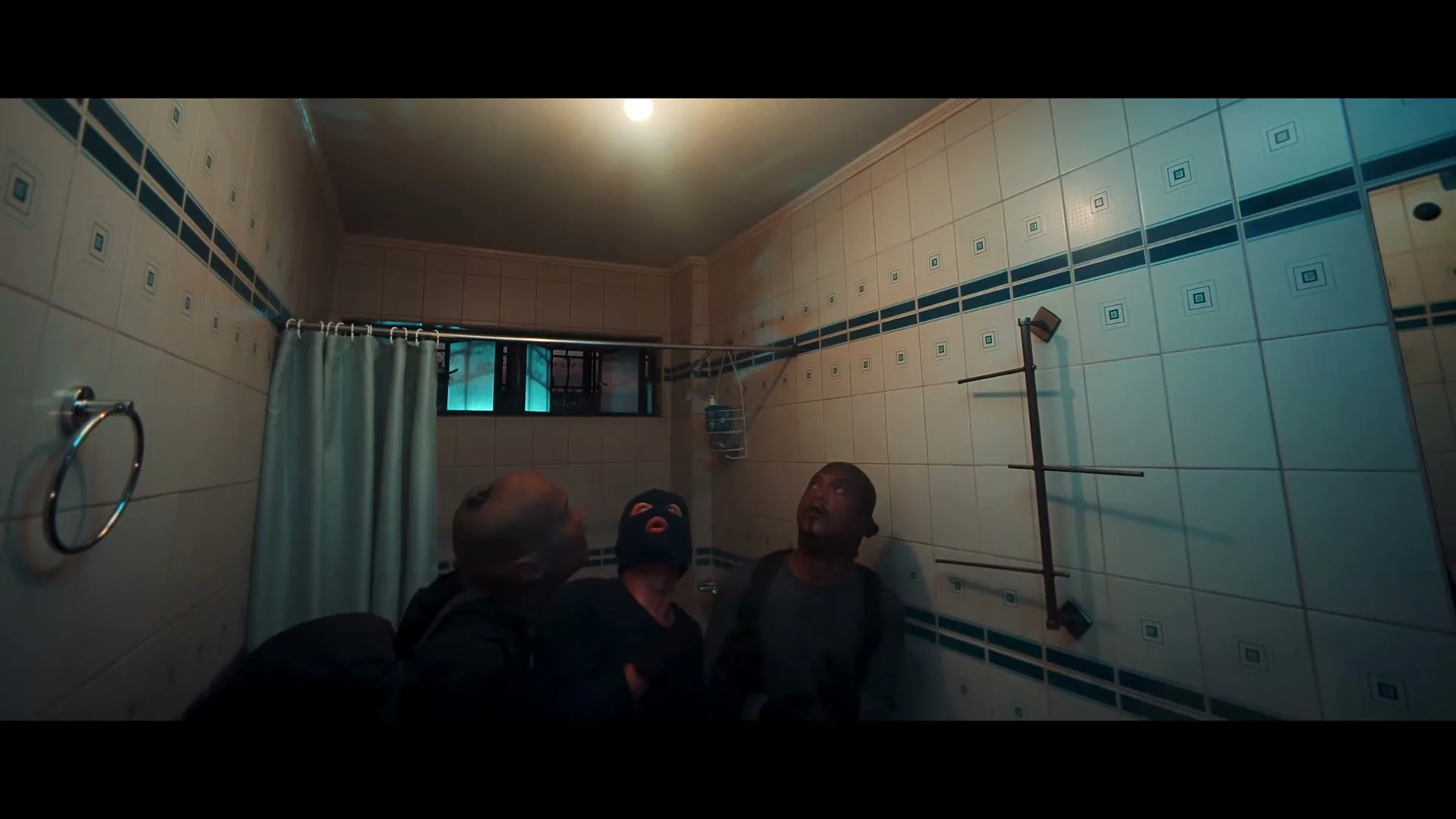 Three men, two with shaved heads and one wearing a black balaclava, look up at a light bulb in a tiled bathroom.