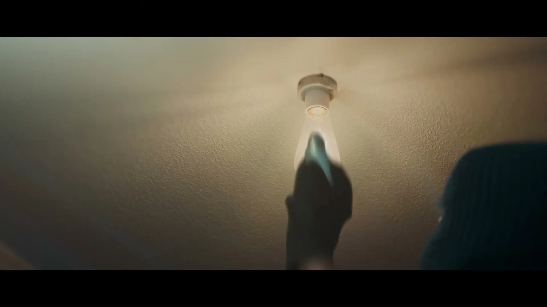 A person in a dark hoodie and gloves screws a lightbulb into a ceiling fixture, illuminating the textured ceiling.