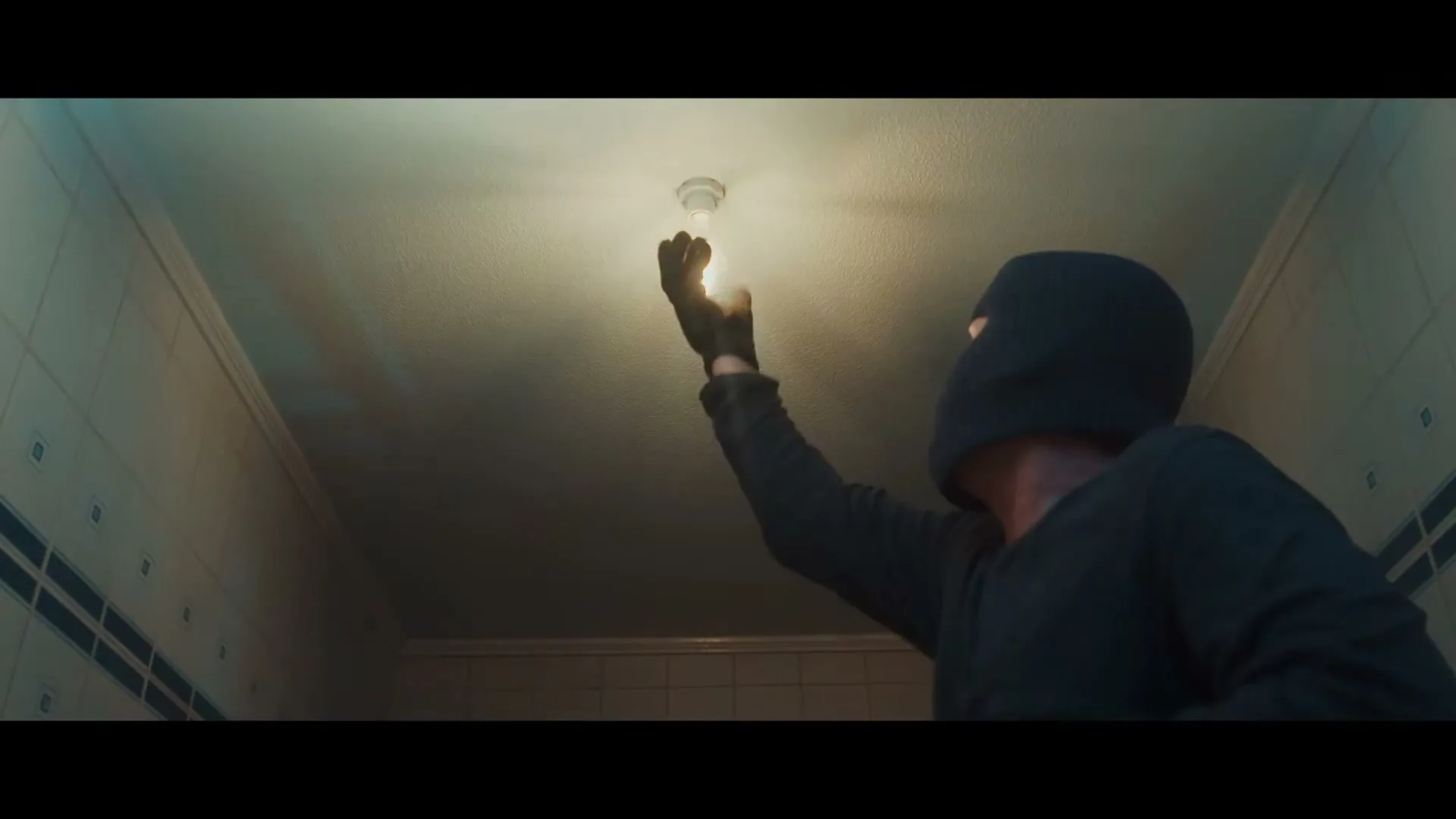 A burglar in a black balaclava and gloves unscrews a lightbulb from the ceiling in a tiled room, stealing it.