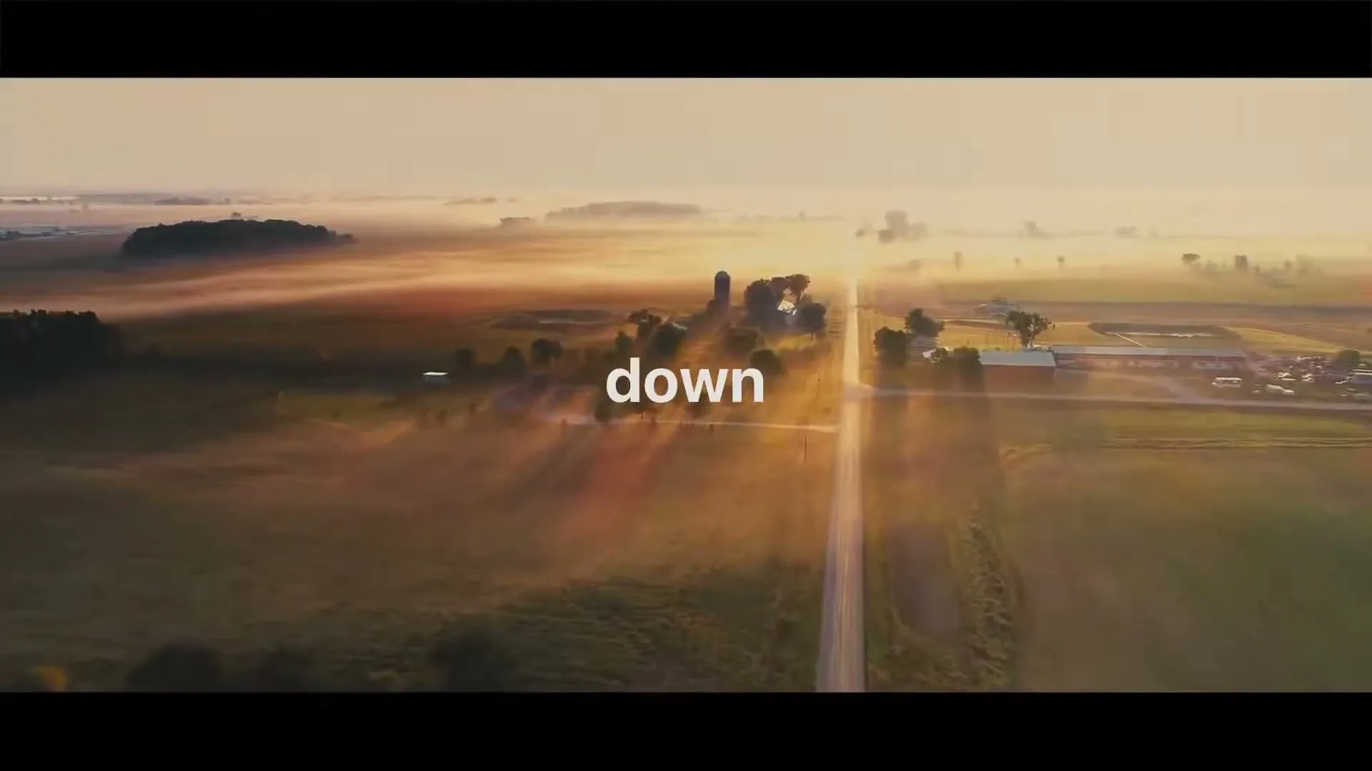 An aerial shot captures a long, straight road cutting through a misty, sun-drenched rural landscape with farms and fields at golden hour.