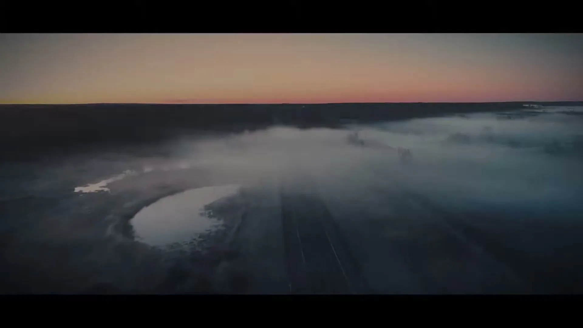 An aerial wide shot captures a misty, dark landscape with a road and a small body of water under a colorful dawn sky.