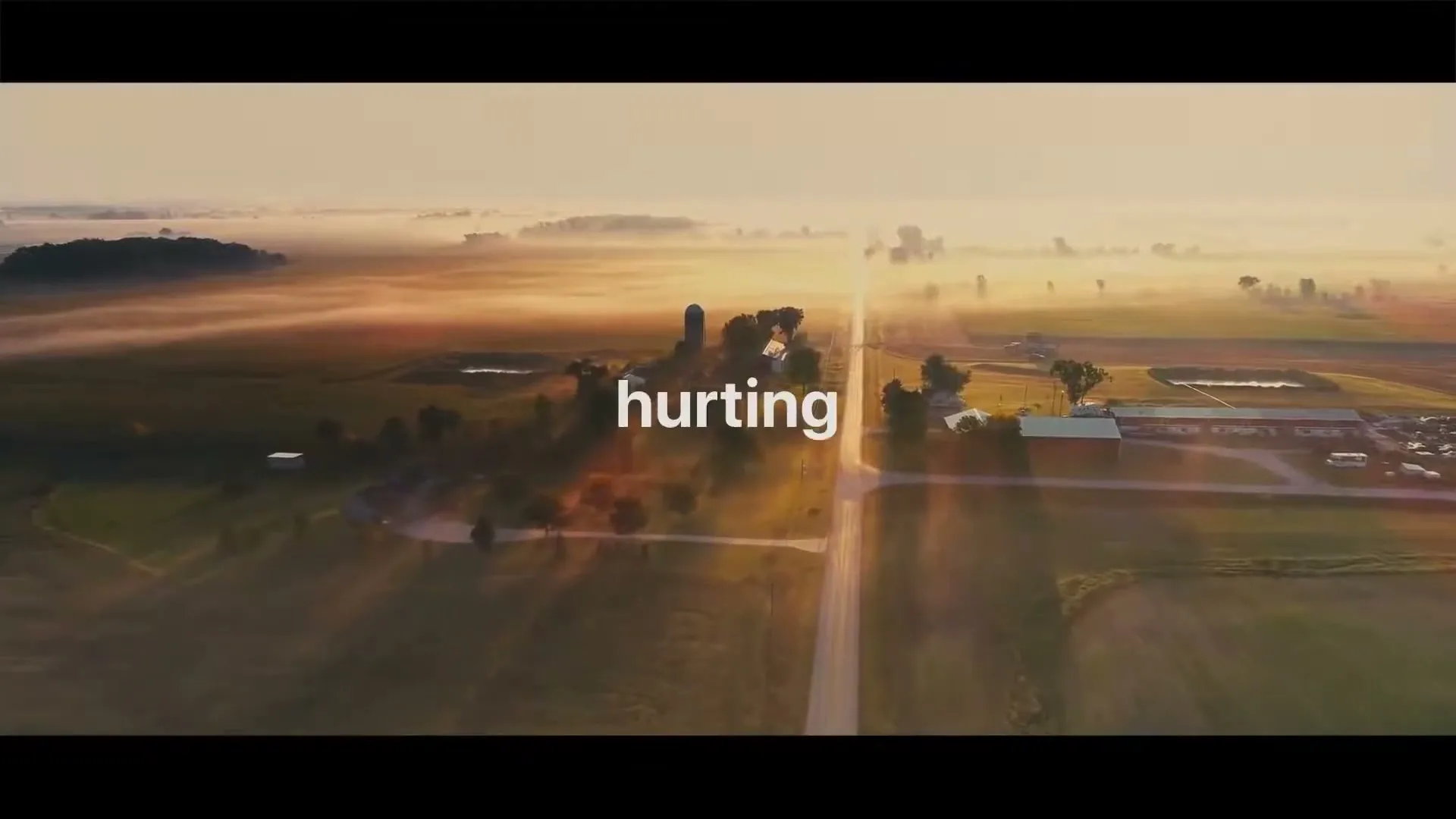 An aerial shot captures a rural landscape with a long road stretching into the distance, surrounded by fields and scattered buildings, under a misty sunrise.
