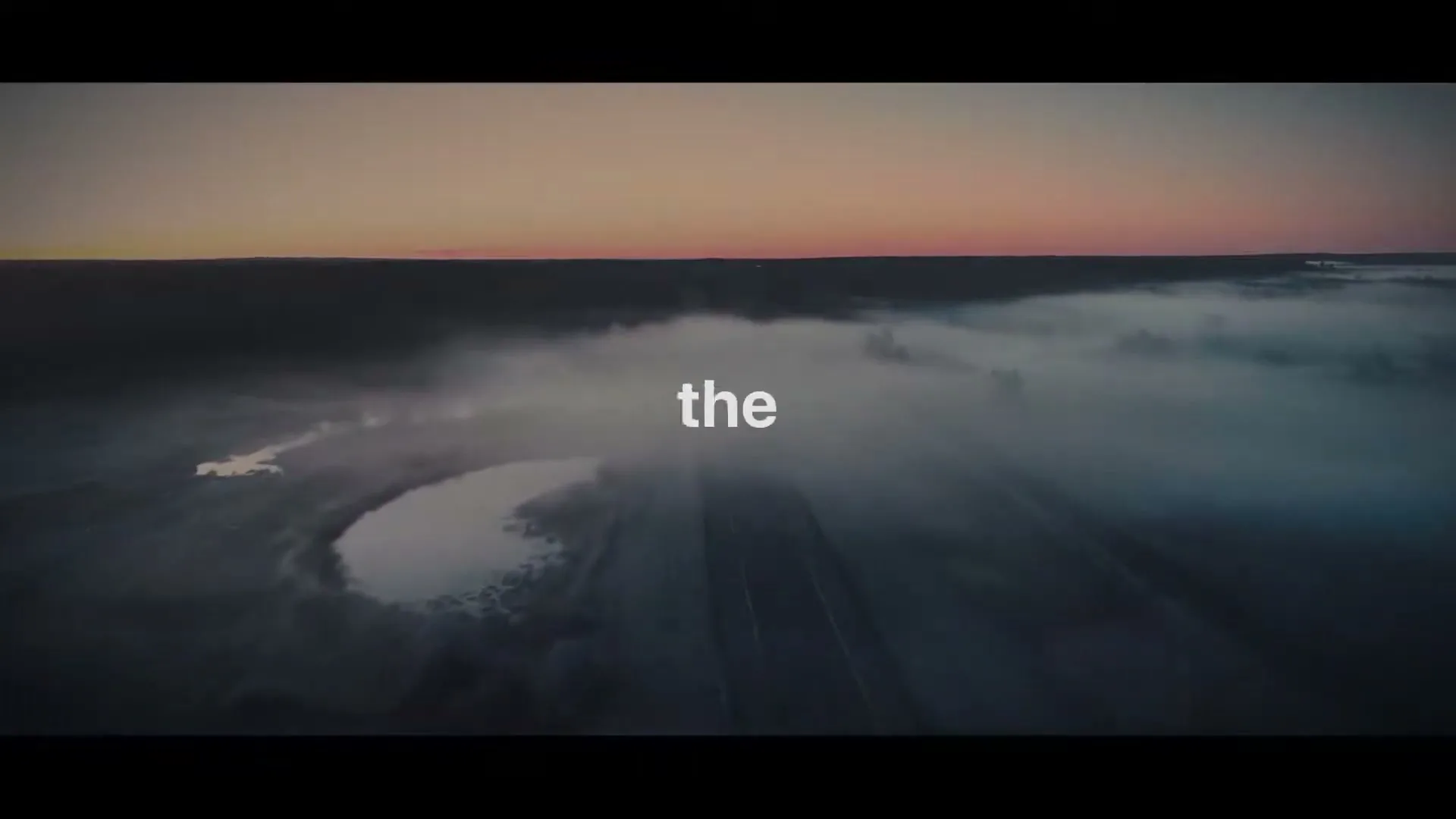 An aerial shot of a vast, misty landscape at dusk, with a road cutting through the fog and small bodies of water reflecting the dim light.