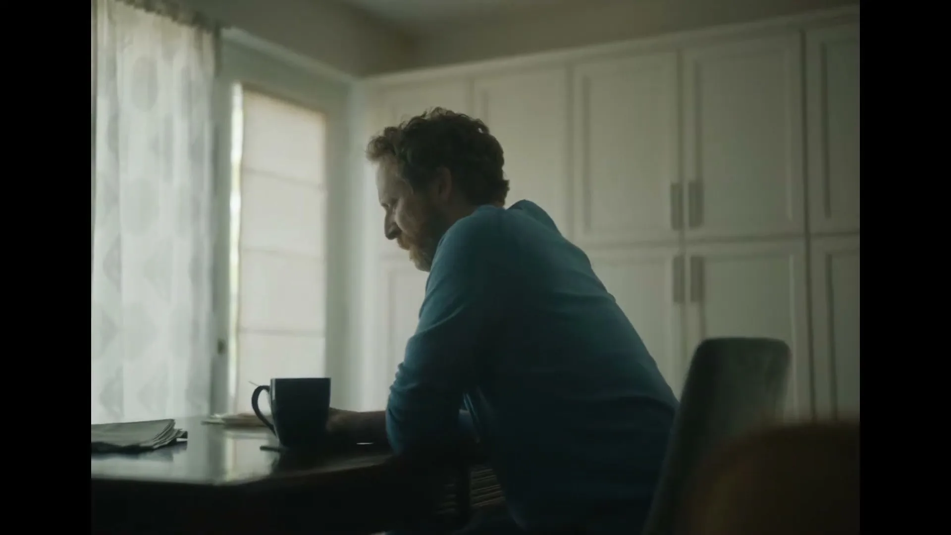 A man with curly red hair and a blue shirt sits at a table with a coffee mug and newspaper, looking down sadly in a kitchen.